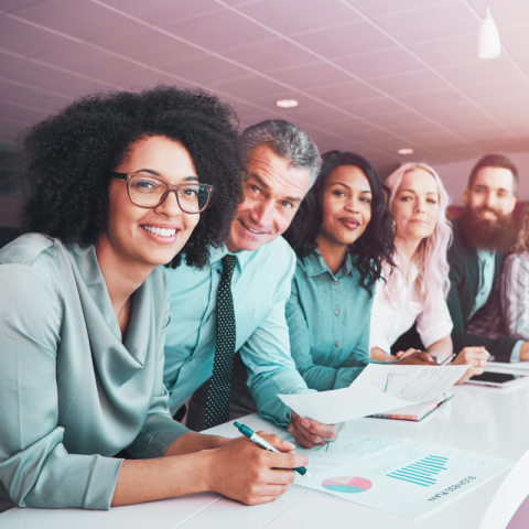 Diverse group of professionals angled along a long, narrow white table that has a few papers with charts and graphs.