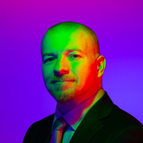 Portrait of Shane Lukas, a middle-aged white man with buzzed hair and goatee in a suit against a black background with multi-colored rainbow lighting