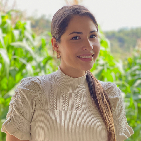 Smiling woman with long dark hair in a light knit top, standing outdoors in front of green plants and a blurred natural background.