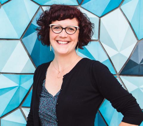 "Smiling woman with short curly auburn hair and cat-eye glasses, wearing a blue top with a black sweater, standing in front of a blue mosaic wall."